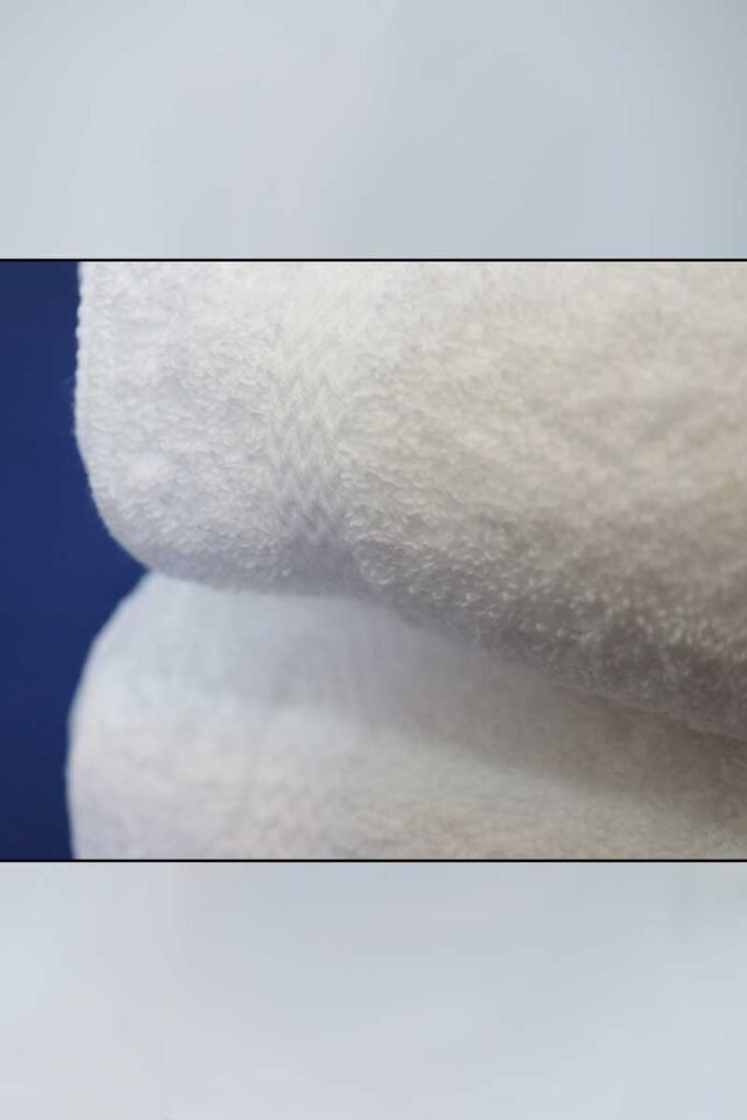 Hotel-Towels-13x13-Wash-Cloth-White - Poltex International Inc Close-up of a white fluffy towel against a blue background, showing soft terry texture and folded edge.